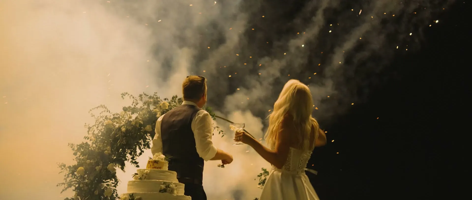 A couple at their cake as sparks rise behind them