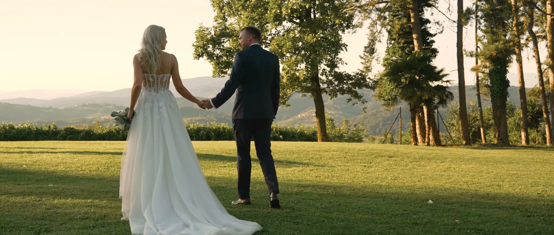 A couple walking hand in hand across a lawn above the vineyards at golden hour